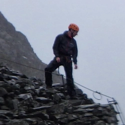 Chris Graham, wearing a helmet and attached to a cable, at the top of a steep rockface